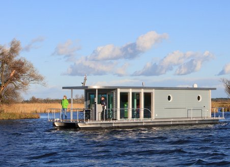 Ein Hausboot schwimmt auf dem Wasser. Es ist grau, zwei Personen stehen auf der Bootsterrasse am vorderen Ende. Im Hintergrund sieht man Felder, Bäume und blauen Himmel mit Wolken.