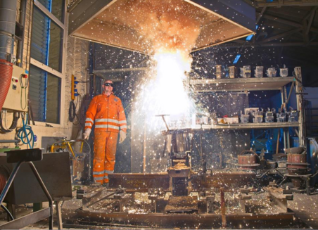 Foto zeigt eine Werkstatt, in der bei einer chemischen Reaktion gelbe Funken fliegen. Ein Mann in Arbeitskleidung und Schutzbrille steht in einiger Entfernung daneben.